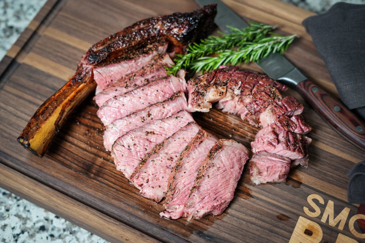 Sliced smoked tomahawk steak fanned out on a wooden cutting board, showing a pink medium-rare interior and charred crust, with the bone intact and garnished with fresh rosemary and a knife in the background.