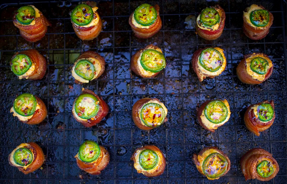 Overhead view of smoked pig shots on a wire rack, featuring crispy bacon-wrapped sausage cups filled with golden cream cheese, topped with sliced jalapeño and sweet BBQ seasoning.